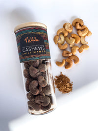 Glass Jar of chili mango chocolate-covered cashews with scattered nuts and fruit pieces on white background
