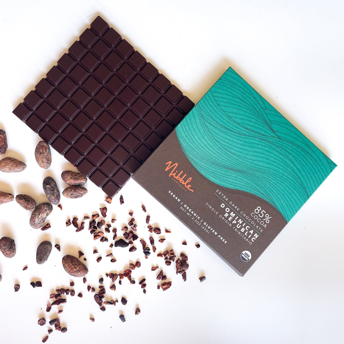 Extra dark chocolate bar with 85% cocoa from the Dominican Republic by Nibble, surrounded by cocoa beans and nibs.