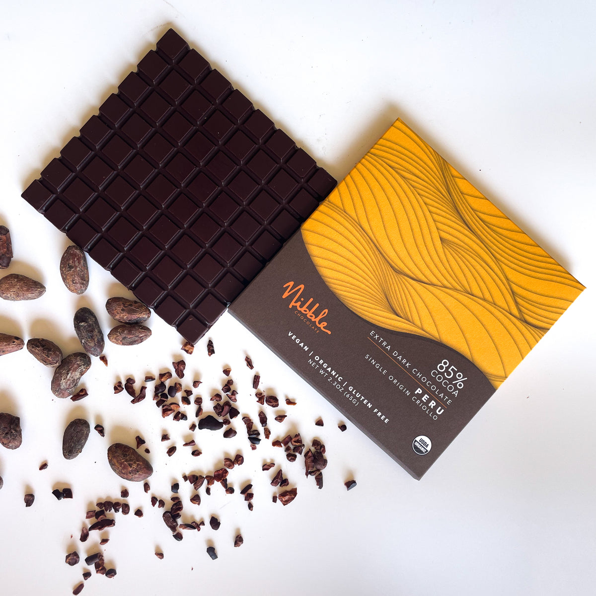 Extra dark chocolate bar with 85% cocoa from Peru, surrounded by cocoa beans and nibs, by Nibble Chocolate.
