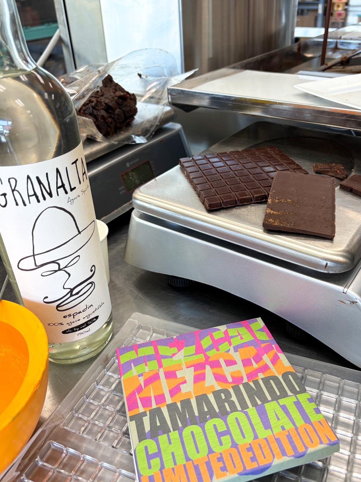 Chocolate bars on a scale with a bottle labeled 'Granalta' and a colorful book titled 'Tamarindo Chocolate White Edition'.