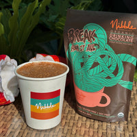 Organic drinking chocolate from Nibble in a cup beside its packaging, set against a tropical background.