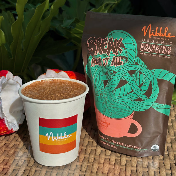 Organic drinking chocolate from Nibble in a cup beside its packaging, set against a tropical background.