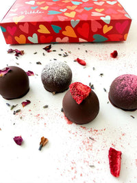 Chocolate truffles with decorative toppings on a white surface, with a colorful heart-patterned box in the background.