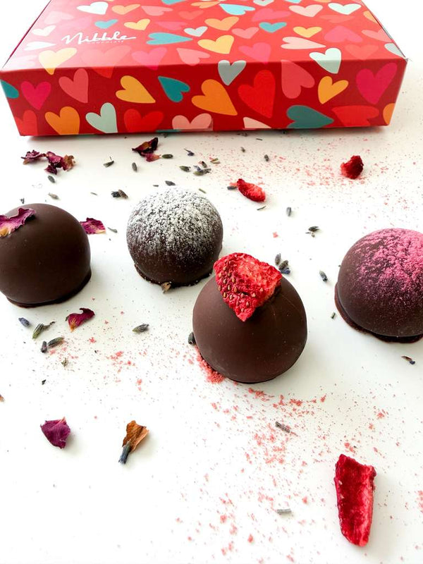 Chocolate truffles with decorative toppings on a white surface, with a colorful heart-patterned box in the background.